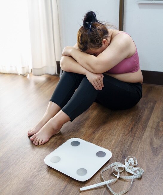 Midlife woman sitting on the floor with head down, scale in the background