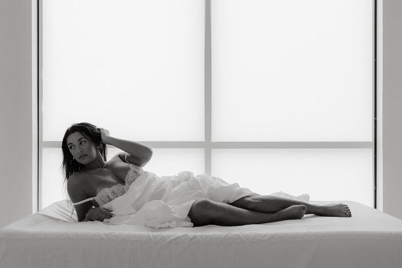 Black and white photo of woman laying on a bed in front of a window wrapped in a white sheet
