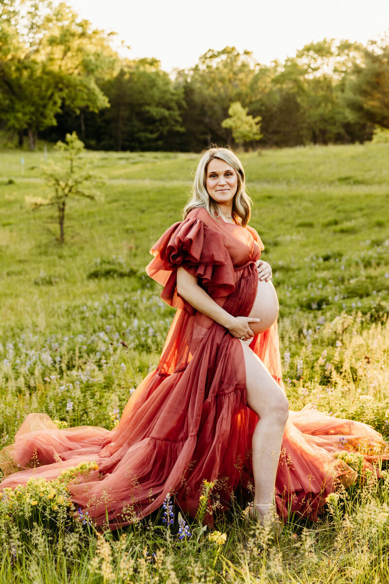 expecting mom holding onto her exposed baby bump thats showing through her coral tulle gown in a field of wildflowers captured by maternity photographer Ashley Kalbus