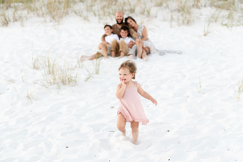Toddler walking ahead of her smiling family during a light-filled beach session in Destin, FL by Andrea Krey Photography