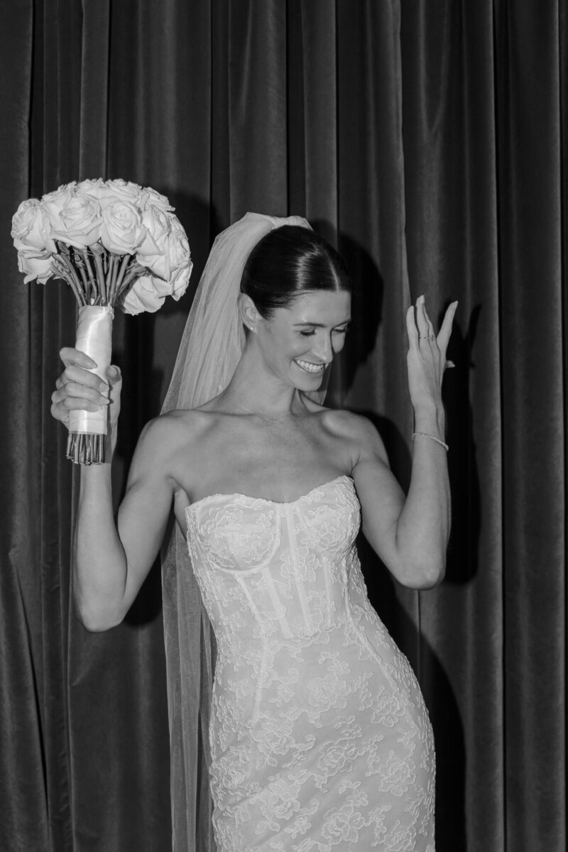 Black and white portrait of a bride with her hands up holding a bouquet in front of a dark curtain