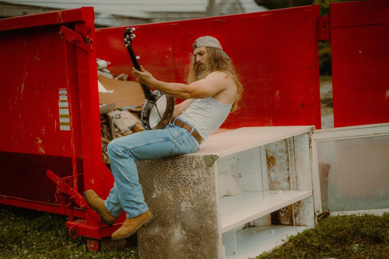 A banjo musician poses in a junkyard for his album cover.