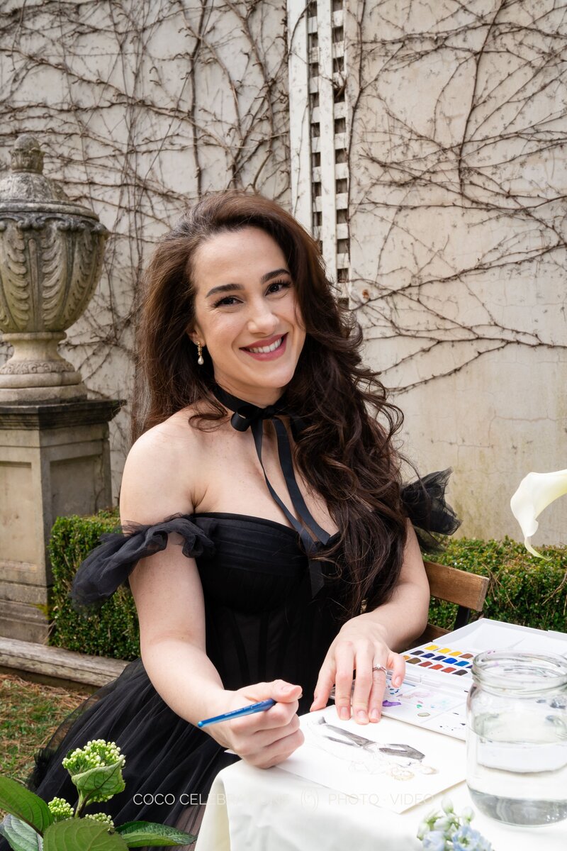 A smiling woman in a black off-shoulder gown sits outdoors at a table, painting live illustrations with watercolors during an event.