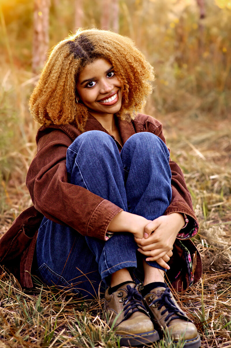 Stunning African American senior girl sitting on grass in a park wearing a brown jacking and jeans in orlando