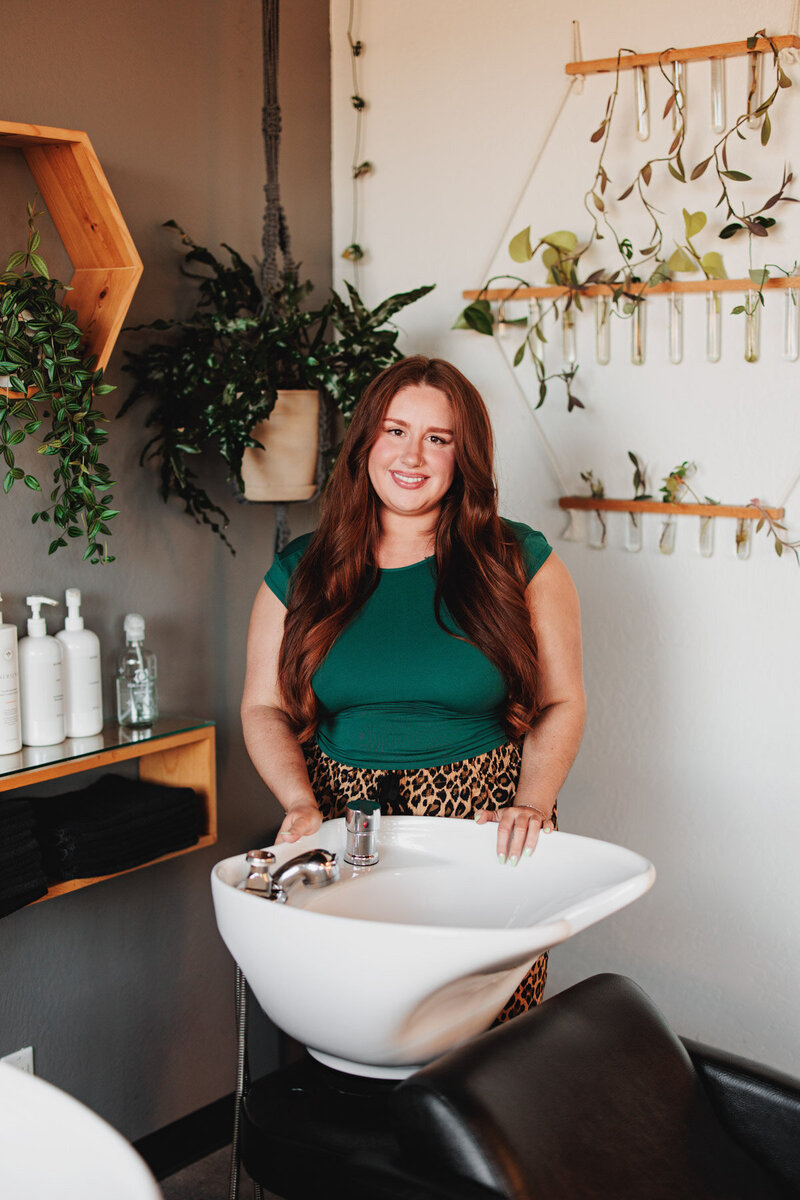 Morgan a hair stylist in Eugene stands behind a shampoo station at the Collective Beauty and Wellness and smiles at the camera.