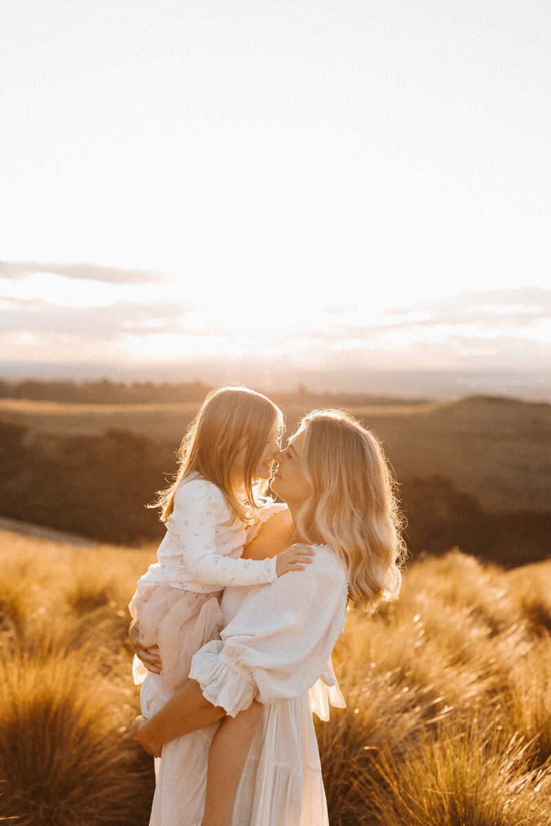 A pregnant mother holding her daughter wearing a white photoshoot dress during her sunset photoshoot with Christchurch maternity photographer lucy jane