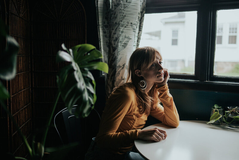 A person in a mustard yellow top sits at a table by a window, resting their head on their hand and looking outside thoughtfully. Nearby, showit designers work on crafting custom Showit website designs. A large leafy plant is in the foreground.