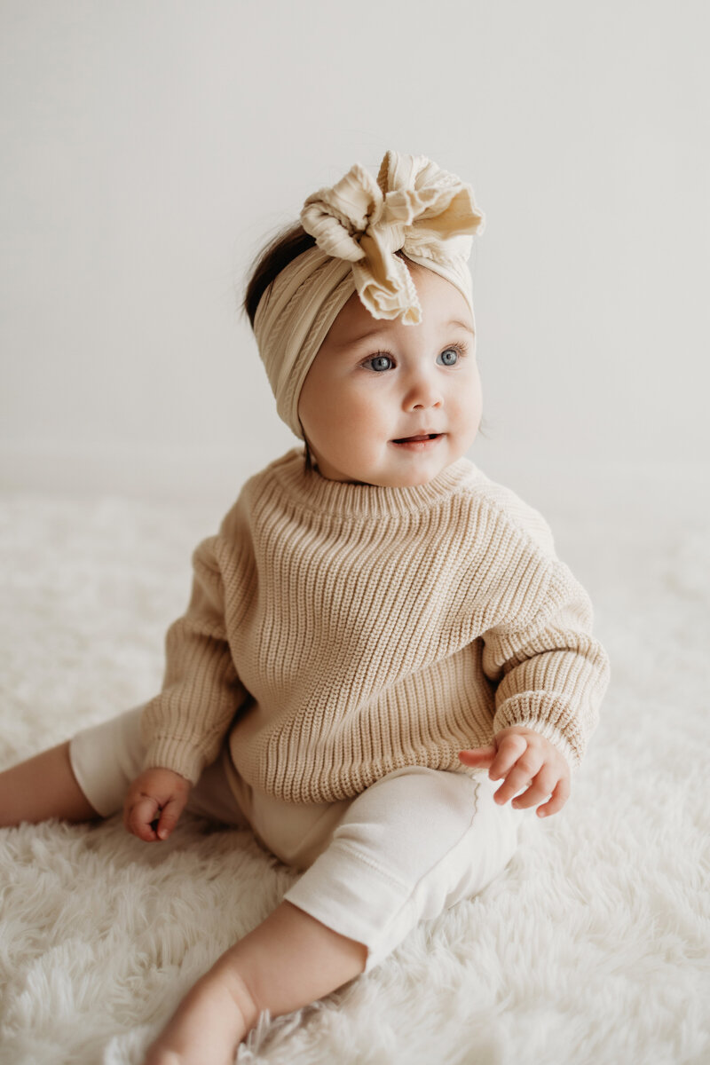 eight month old her girl dressed in a cream knit sweater and a large cream bow stares out the window during her Denver milestone session