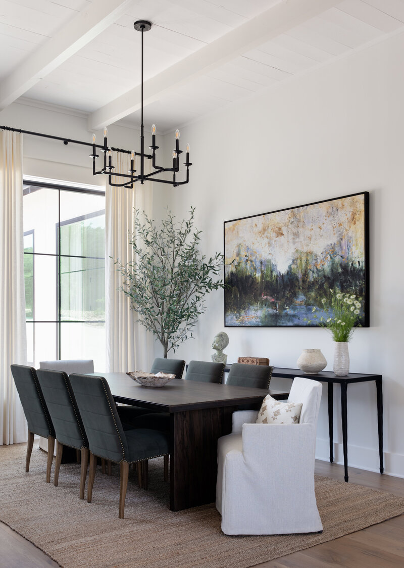 Dark wooden table on jute rug with blue upholstered chairs. Colorful art on white walls with large windows looking outside.