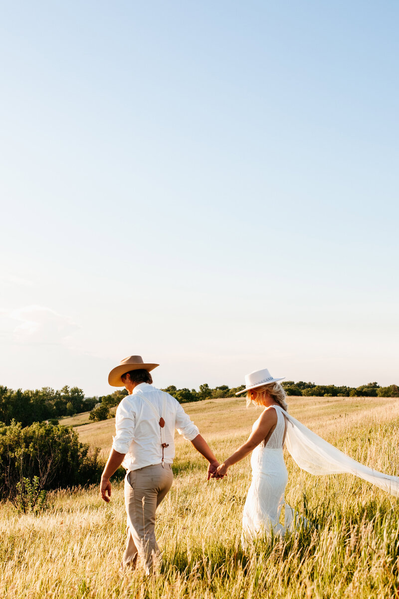 Groom in khaki pants and matching cowboy hat leads his wife in a white dress and cowboy hat as they walk down a grassy hill in front of Minneapolis wedding photographer