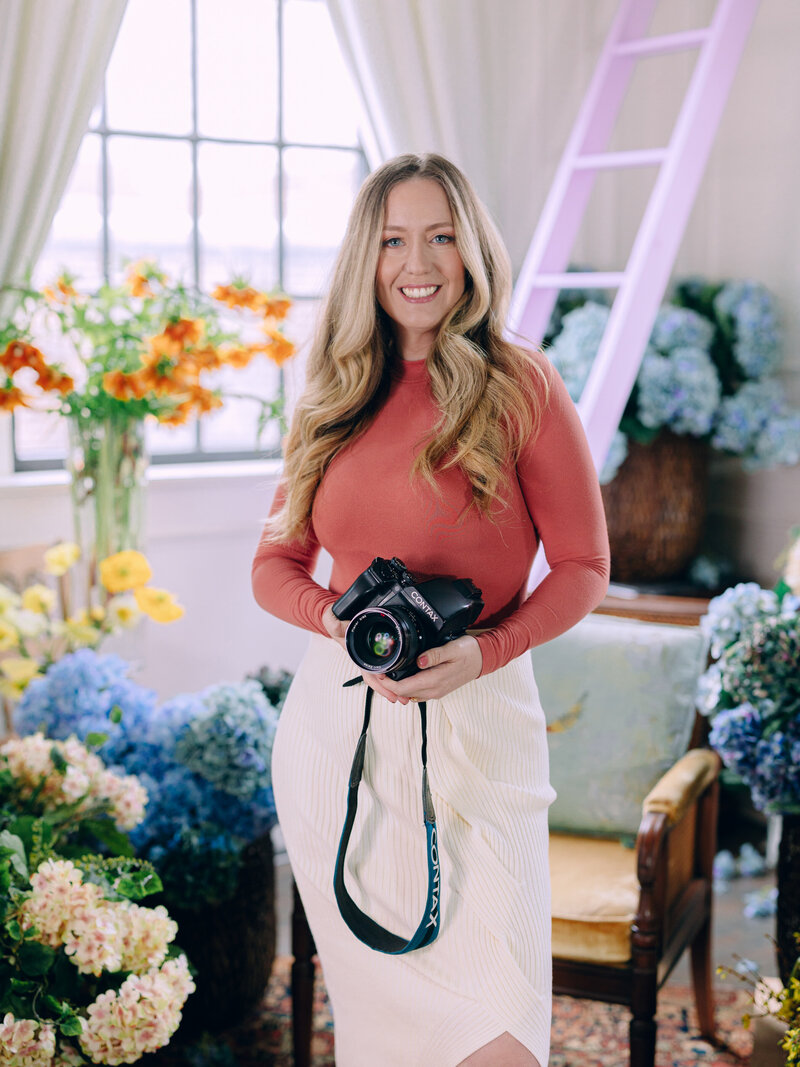 headshot of a woman holding a film camera surrounded by buckets of flowers