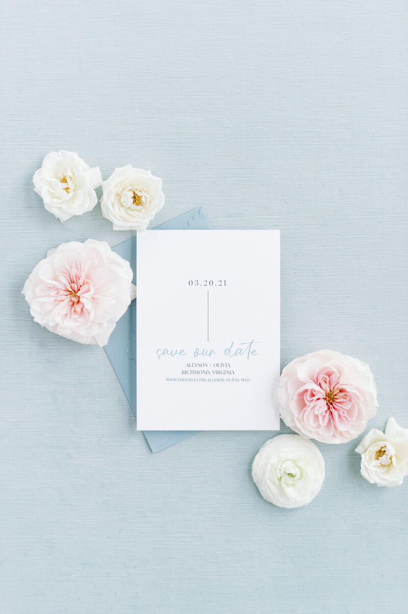 Dusty blue save the date card displayed on a blue styling mat with flowers.