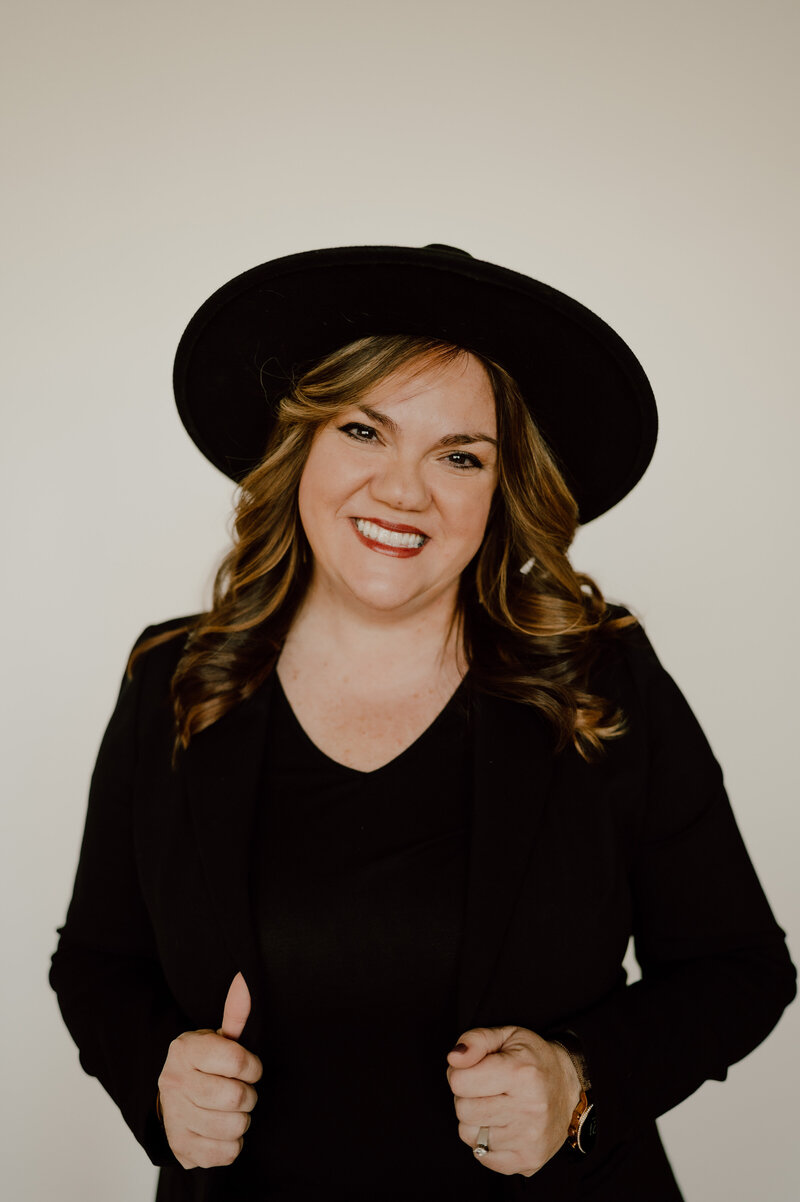 Woman in black blazer and hat, who is a wedding planner from Illinois, smiling at the camera with a plain light background.