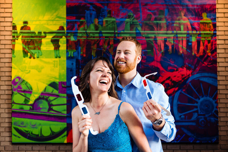 A couple holding 3d glasses laughs in front of a  3d mural in downtown Durham, NC