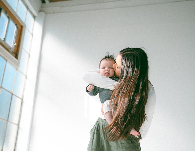 Mother holding and kissing her newborn baby in soft window light during a lifestyle family photography session in Orange County.