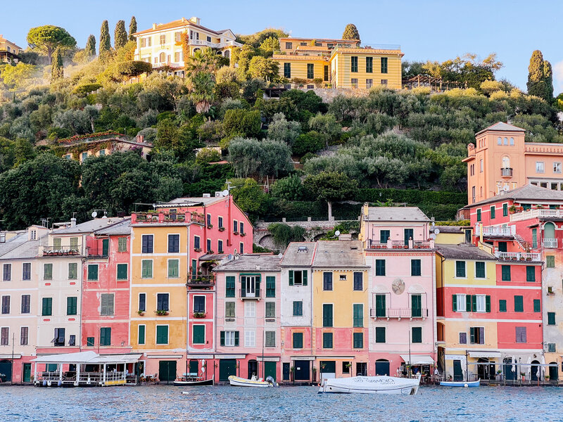 Colorful buildings line the waterfront in a picturesque coastal village, with lush green hills and villas in the background. Boats are anchored in the calm, blue water.