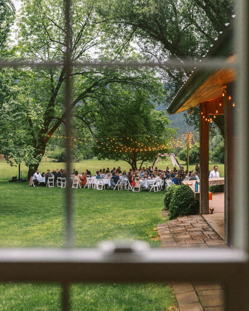 A wedding reception with stringed lights and guests seen through a window pane, representing the style of Oregon wedding photographer Among the Oaks. 