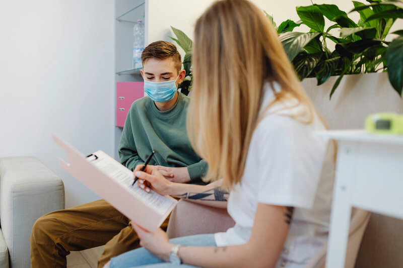 Teenager wearing a protective face mask sitting with a healthcare worker who is holding a clipboard and pen.