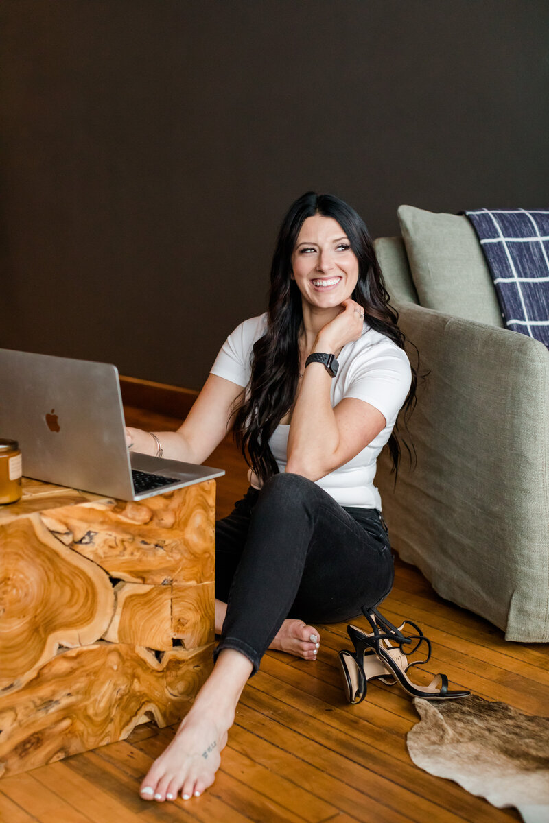 Allie Clark seated on the floor beside a modern wood coffee table, smiling while working on her MacBook Pro—capturing the approachable, intentional energy behind Allie Clark Designs.