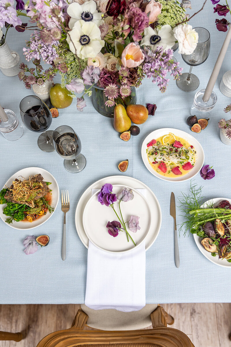 Event table setting with gourmet food plated. The table is decorated with tinted wine glasses, purple flowers, and pears and figs