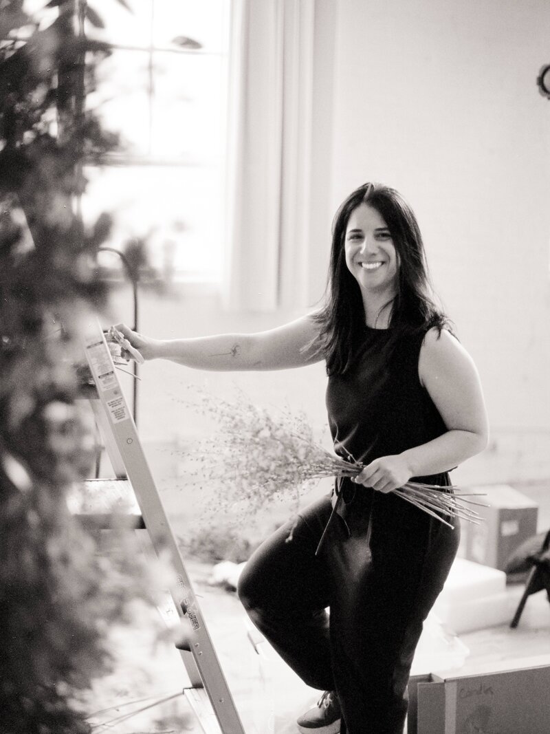 Black-and-white photo of florist smiling at the camera while holding delicate filler stems in one hand and steadying a ladder with the other in a bright studio.