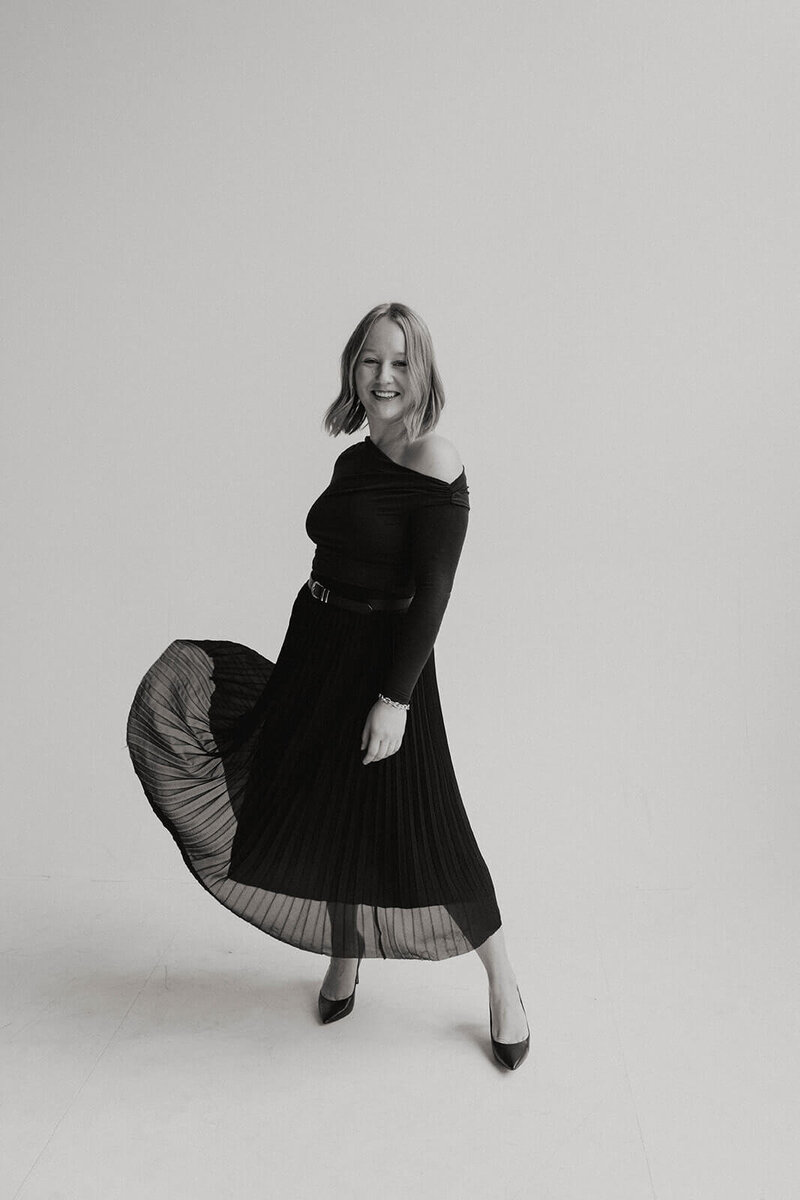 A woman posing in a flowy black dress smiling at the camera on a white backdrop