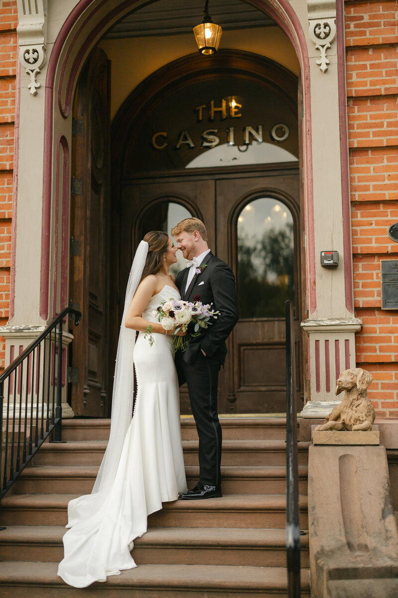 A bride and groom share an intimate moment under a veil. The groom, wearing a suit, kisses the brides cheek. The bride wears a strapless dress and smiles softly with her eyes closed. The black-and-white image creates a timeless, romantic feel.