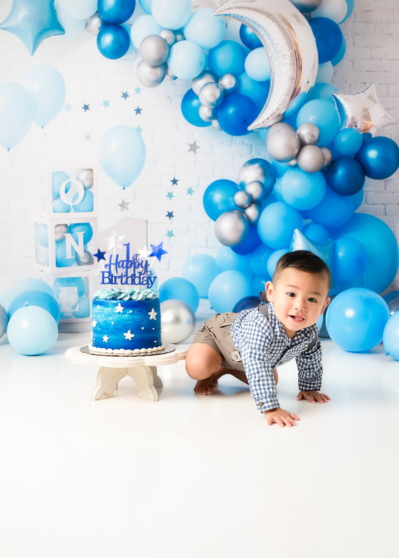Cake smash photography — baby boy with birthday cake and moon-themed blue balloon garland for first birthday celebration