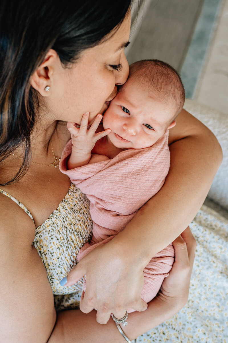 A tender moment between a mother and her newborn baby, wrapped in a pink blanket. The mother is gently kissing the baby’s forehead while holding them close.