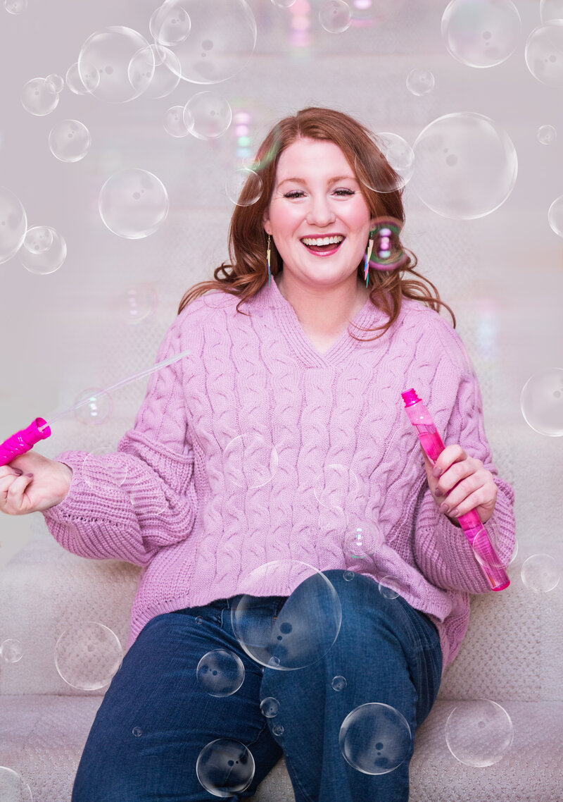 A redheaded woman in her mid-twenties sits on the steps in a lilac sweater and jeans and smiles at the camera with bubbles surrounding her.
