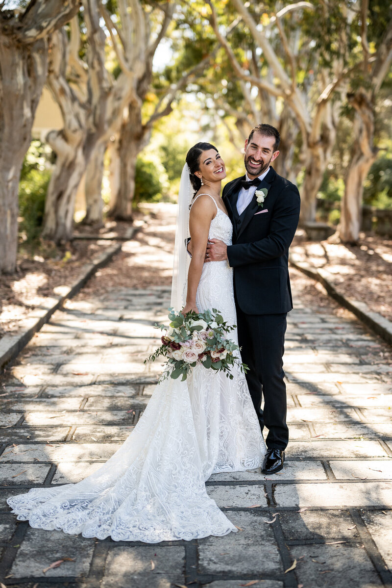 Professional San Francisco Bay Area wedding photography featuring authentic moment as bride in white lace mermaid dress with veil and groom in black tuxedo share genuine laughter on stone pathway under natural tree canopy with dappled sunlight creating romantic outdoor atmosphere