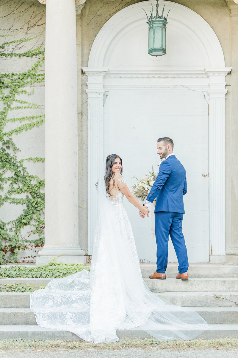 Bride poses for an editorial-style bridal portrait at Chateau des Champlatreux in Paris, France. Her off-the shoulder gown features sheer fabric, boning, and is embroidered with floral details. Her brown hair is tied back and she is looking over her shoulder with a closed-mouth smile. Captured by top destination wedding photographer Lia Rose Weddings