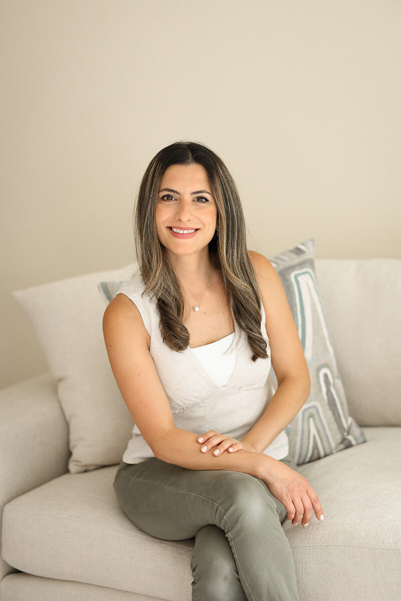A woman, Katia Mashni, a nutritionist, sits on a white couch with her hands resting on her knees, looking thoughtful.