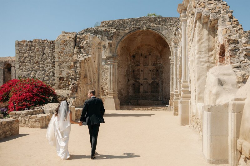 Photo from behind of bride and groom holding hands and walking towards a large old stone structure