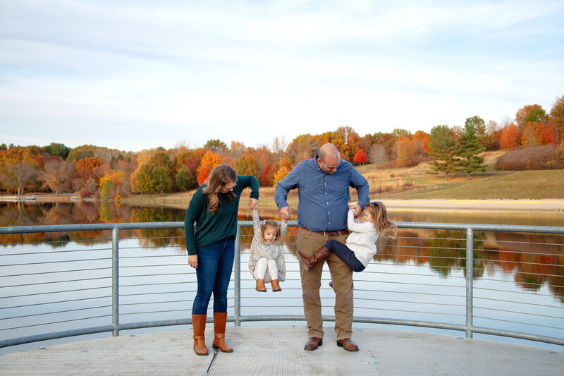 Mom and Dad lifting their two young girls in front of lake and colorful fall trees.