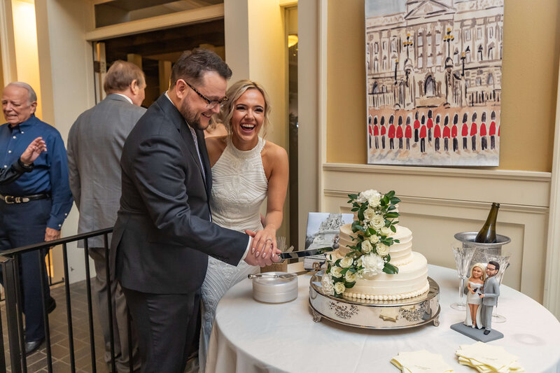 A smiling bride and groom cut a white, floral wedding cake together at a reception.