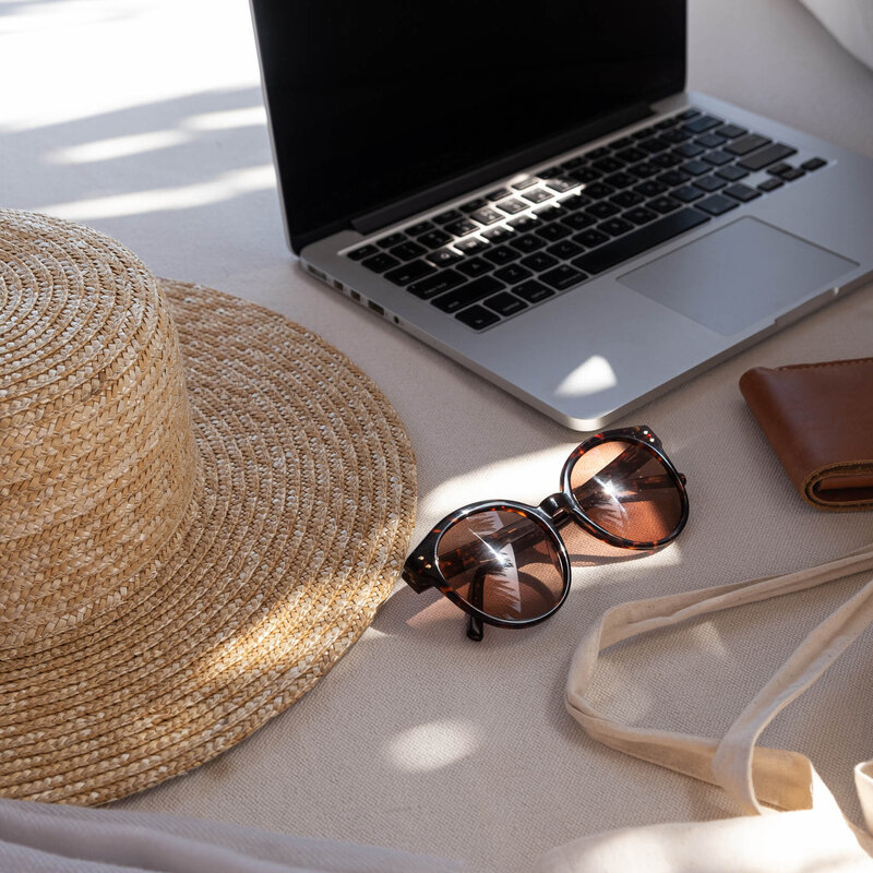 A straw hat, a pair of round sunglasses, a laptop, and a brown wallet on a light-colored surface with soft sunlight and shadows.