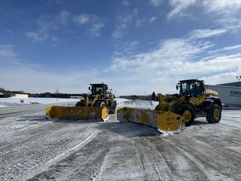 Two modern yellow snowplows positioned on a snow-covered surface under clear blue skies