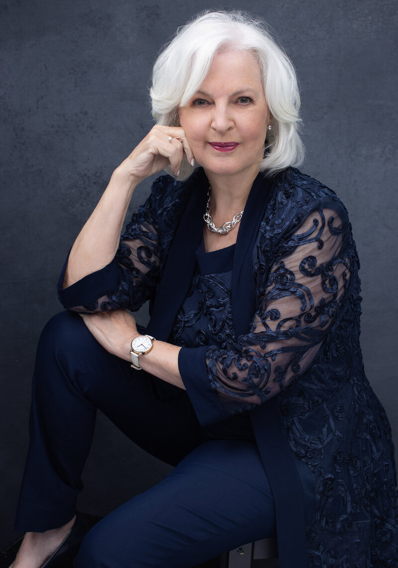 Elegant senior woman with striking white hair, styled in a sophisticated updo, confidently poses at a Cincinnati portrait studio. Dressed in a chic navy ensemble complemented by intricate lace detailing and a bold silver necklace, her poised expression exudes grace and experience.