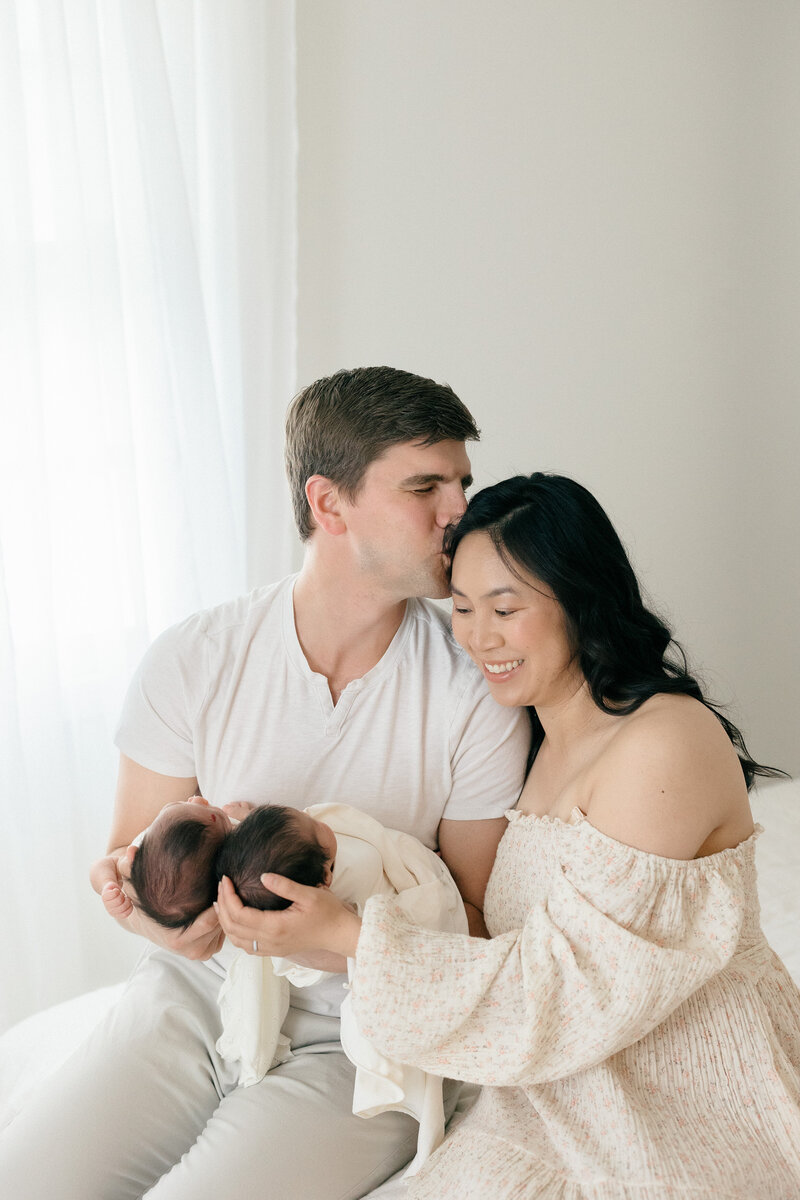 A dad kissing his wife's temple while holding newborn twins in all white studio by New Jersey baby photographer