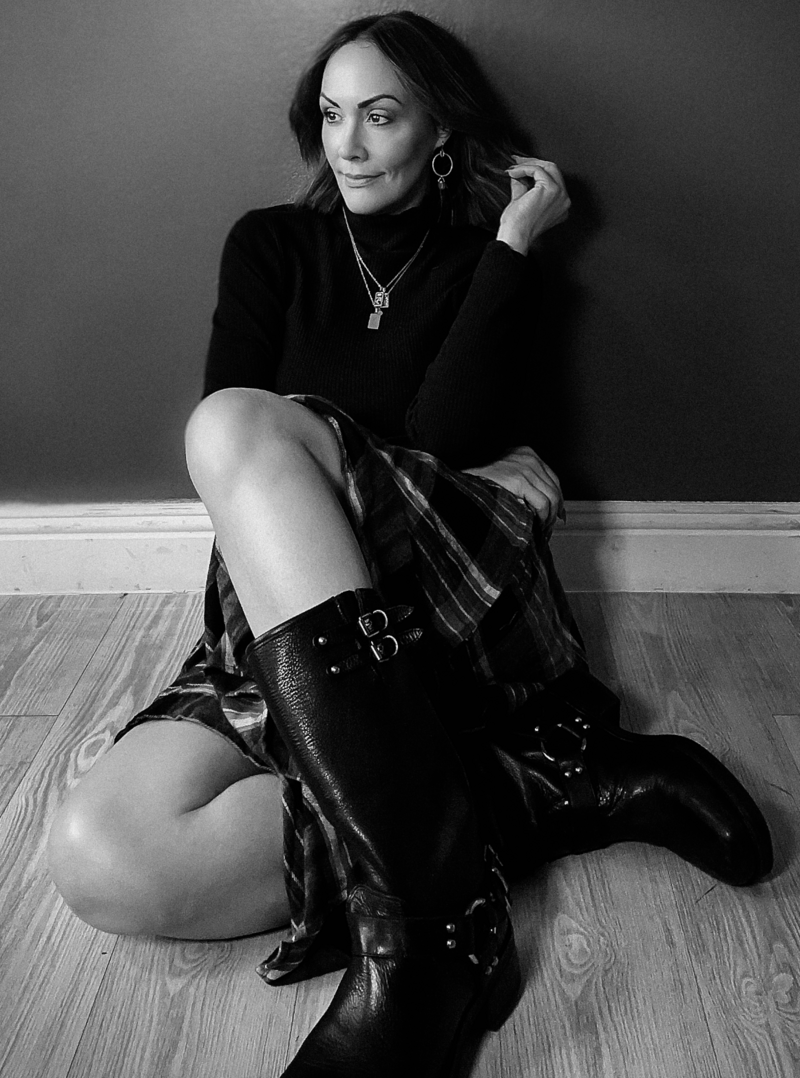 black and white photo of a woman sitting down leaning against a wall looking off to the side she has short hair and black biker boots on