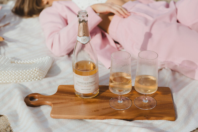 A bottle of Crémant de Bordeaux Brut Rosé and two filled glasses on a wooden tray, with a person in a pink outfit in the background. The setting is a casual outdoor picnic. The text overlay reads 'Social Hang,' emphasizing Shindig Social's focus on fostering social gatherings.