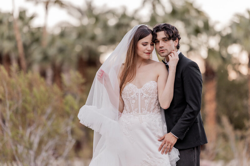 A striking bride and groom pose beside a vintage convertible in the Palm Springs desert, exuding timeless elegance and cinematic romance. The bride’s ethereal veil flows in the breeze, while the groom’s sharp black tuxedo adds a classic touch to this sophisticated wedding portrait.