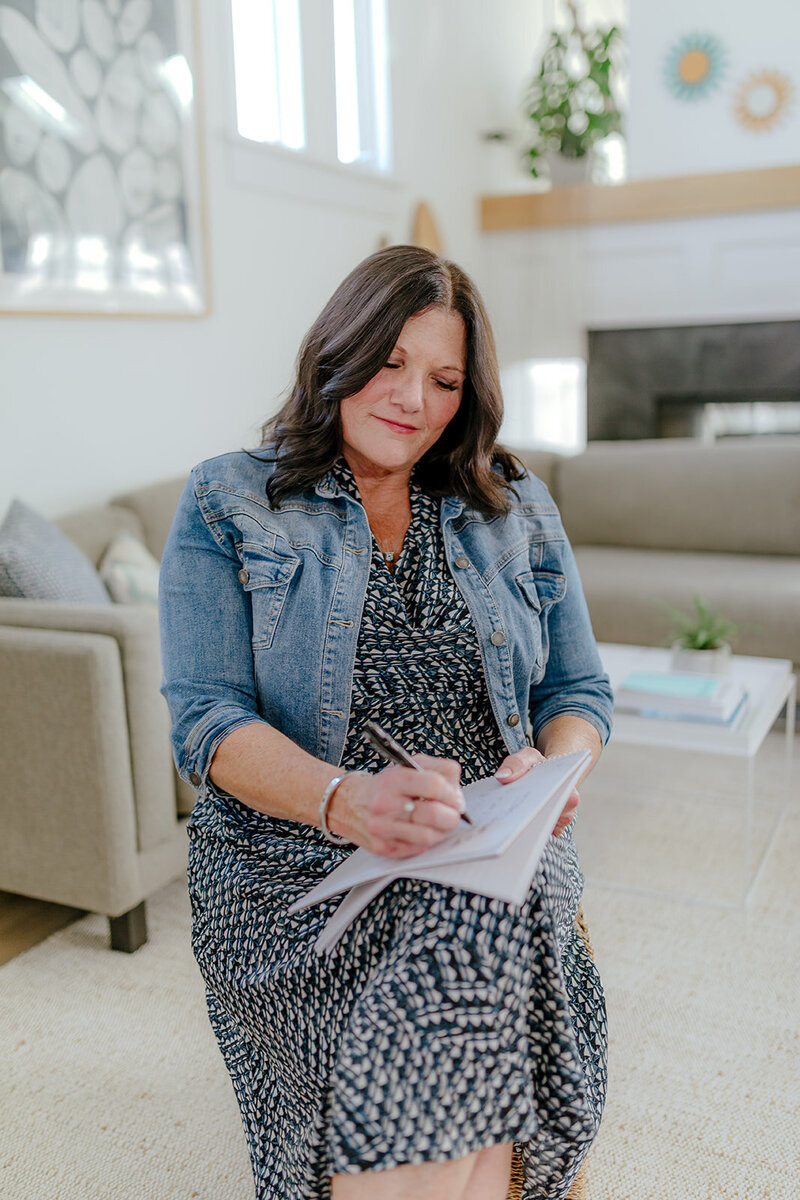 Susie Schumacher writing in a notebook while seated in a comfortable, modern living room, wearing a denim jacket and patterned dress, focused and reflective - Susie Schumacher Life Coach