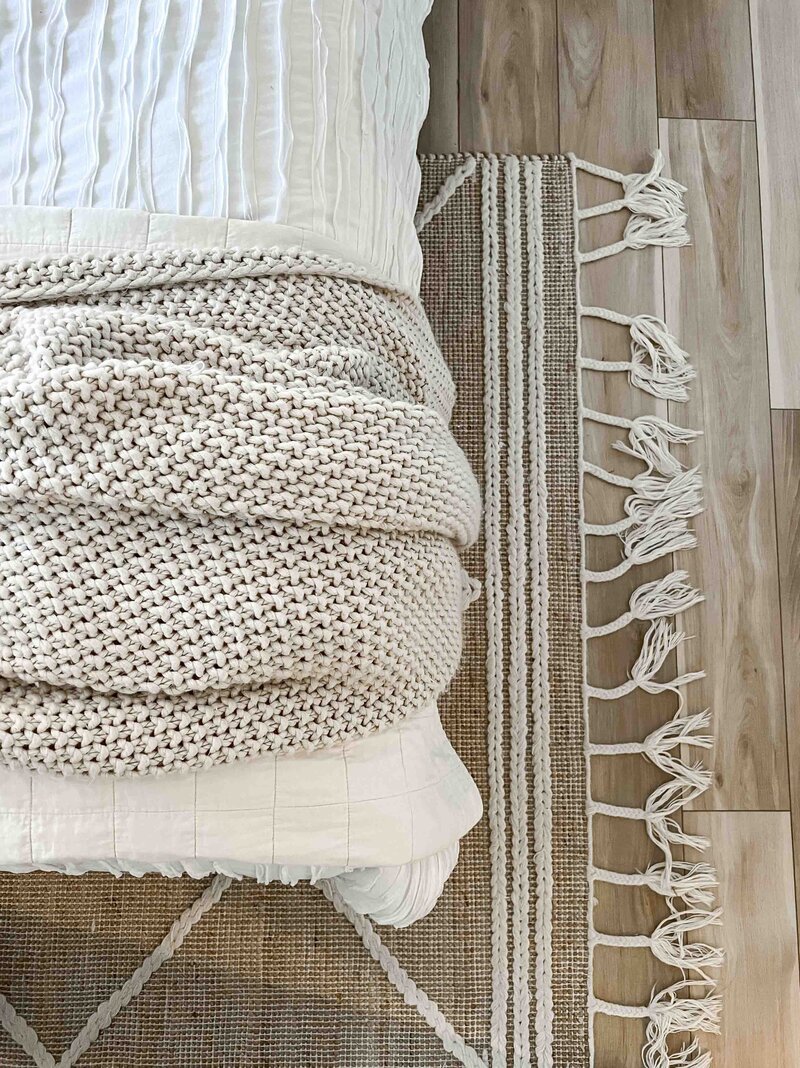 top view of wood floors, boho neutral rug with white tassels, bed with white bedding and neutral throw blanket on the end