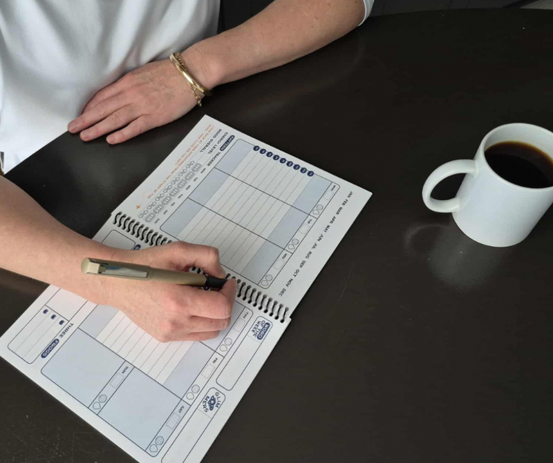 A person writing in the weekly spread of the Time Magic Planner with a pen, seated at a dark table with a cup of coffee nearby.