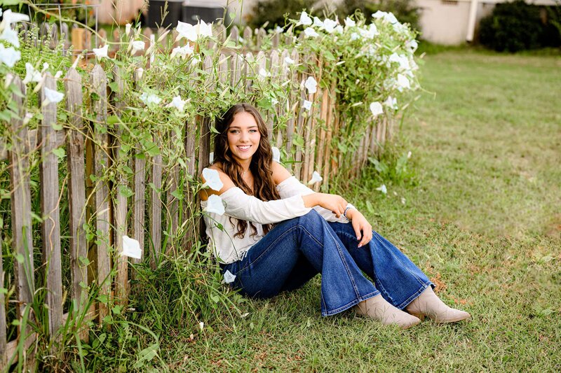 wearing jeans and a white top leaning against a fenceline covered in blooms