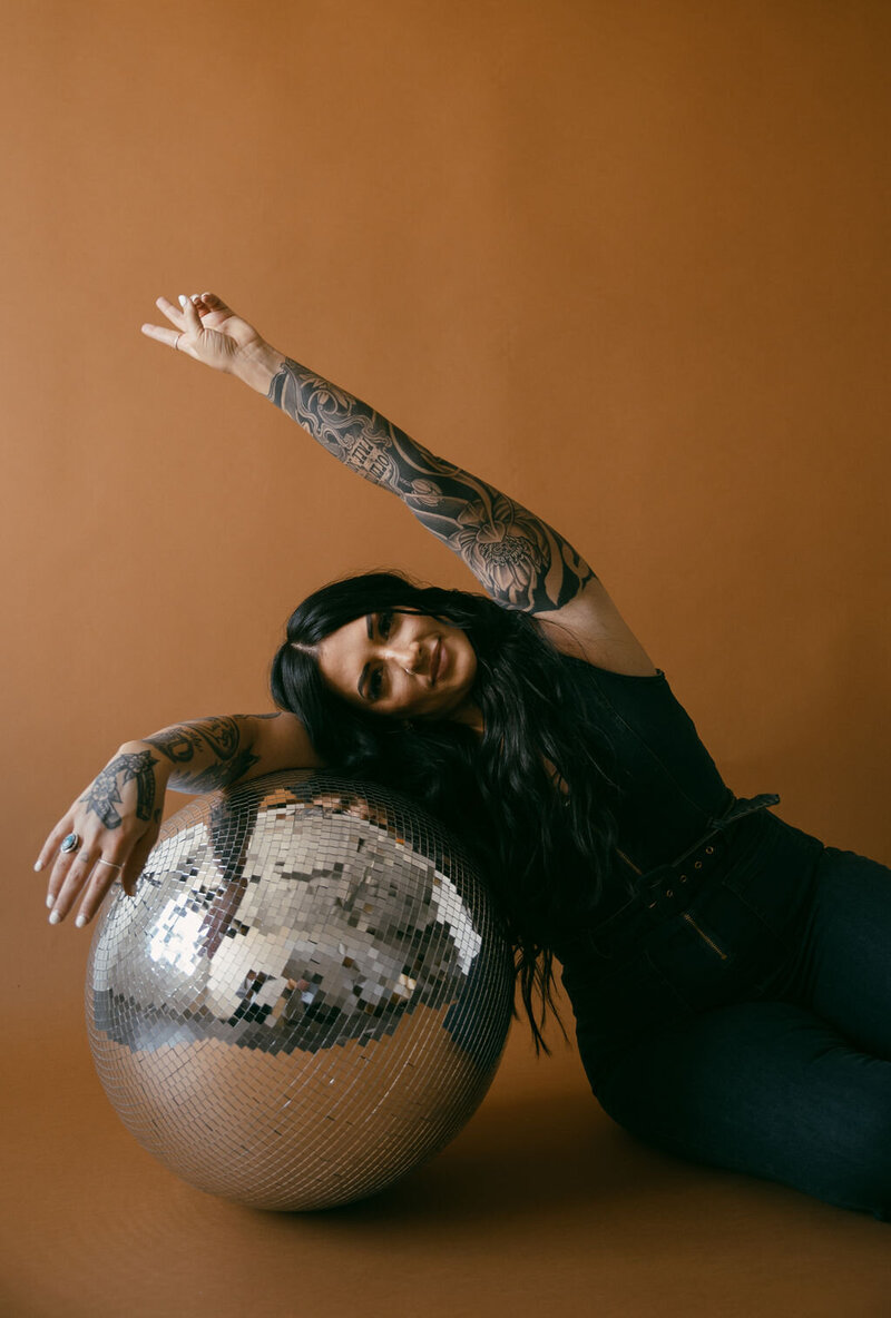 tattooed woman leaning against a large disco ball, smiling