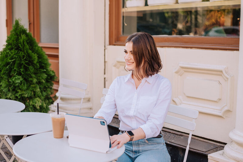 Madison smiles while working on her iPad at an outdoor café table, representing her approachable and professional vibe as a creative partner for women entrepreneurs.