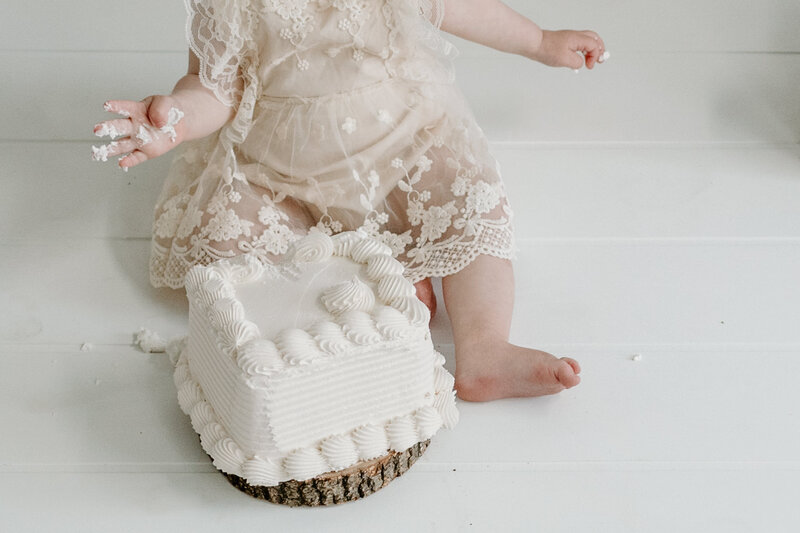 Cake smash photo of white cake with baby girl sitting behind cake in white lace dress.